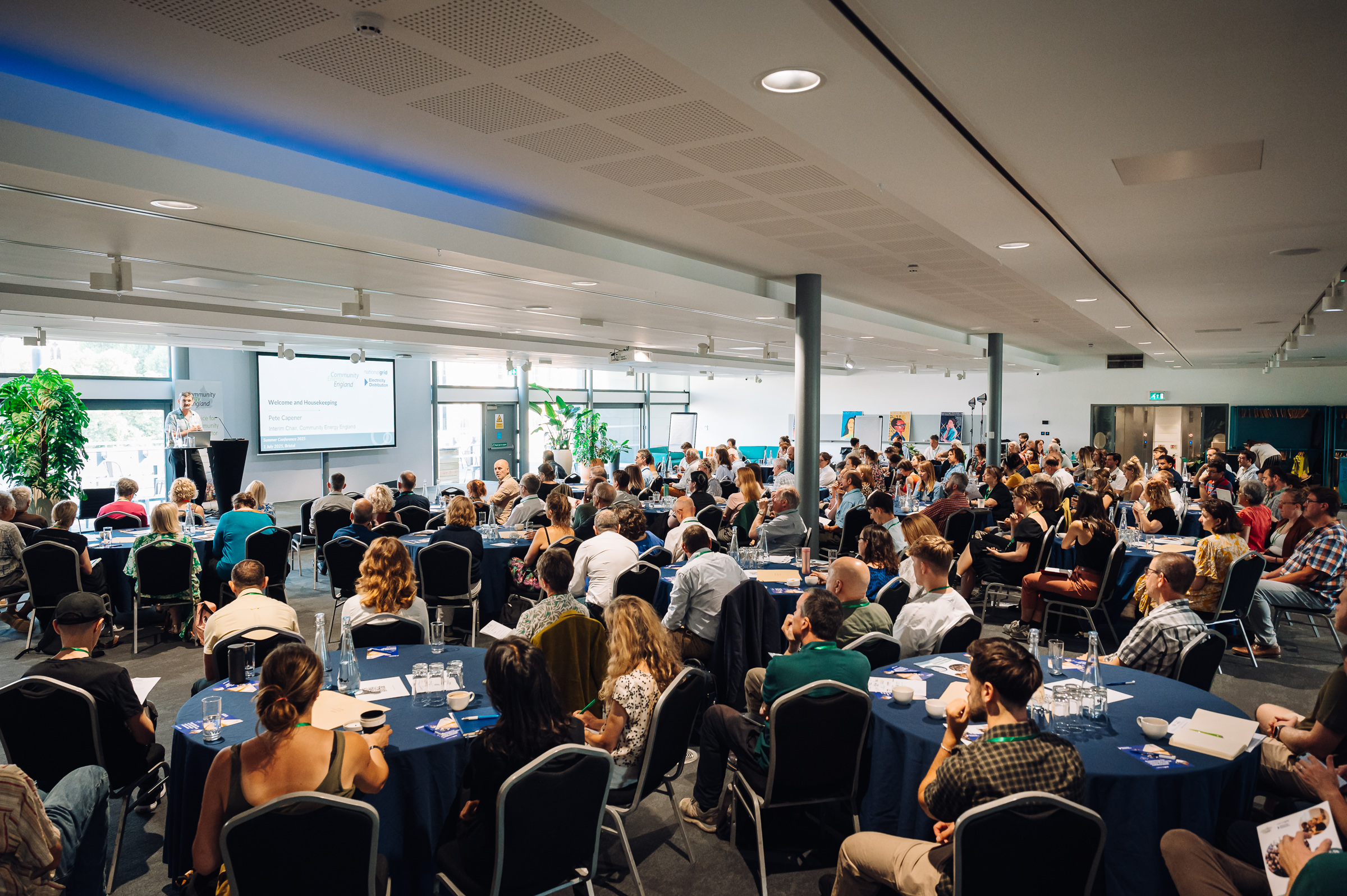 a large room filled with people sitting at round tables, all looking forward at a speaker standing at a podium on a stage