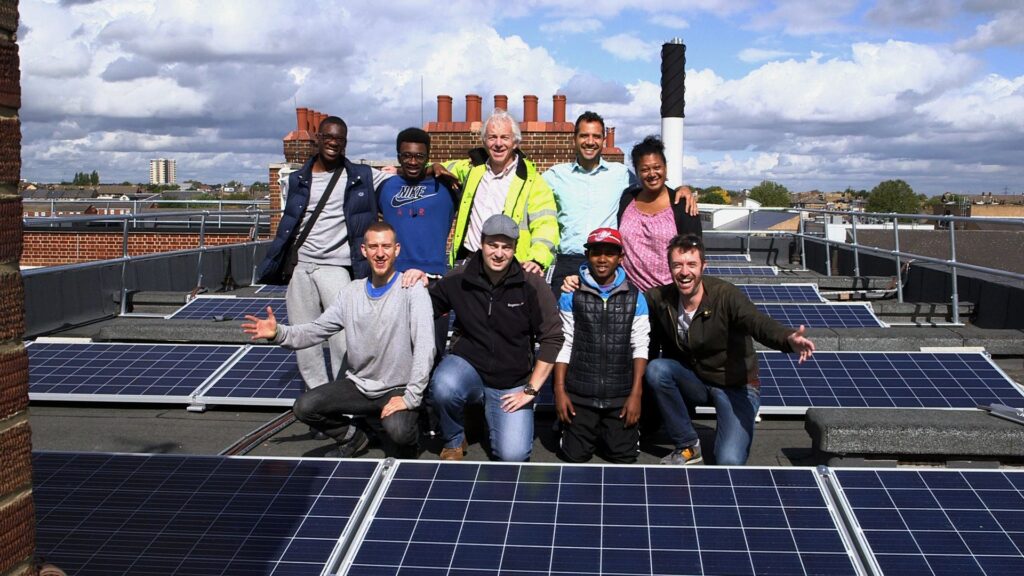 Nine smiling people stand on a roof with solar panels all around them