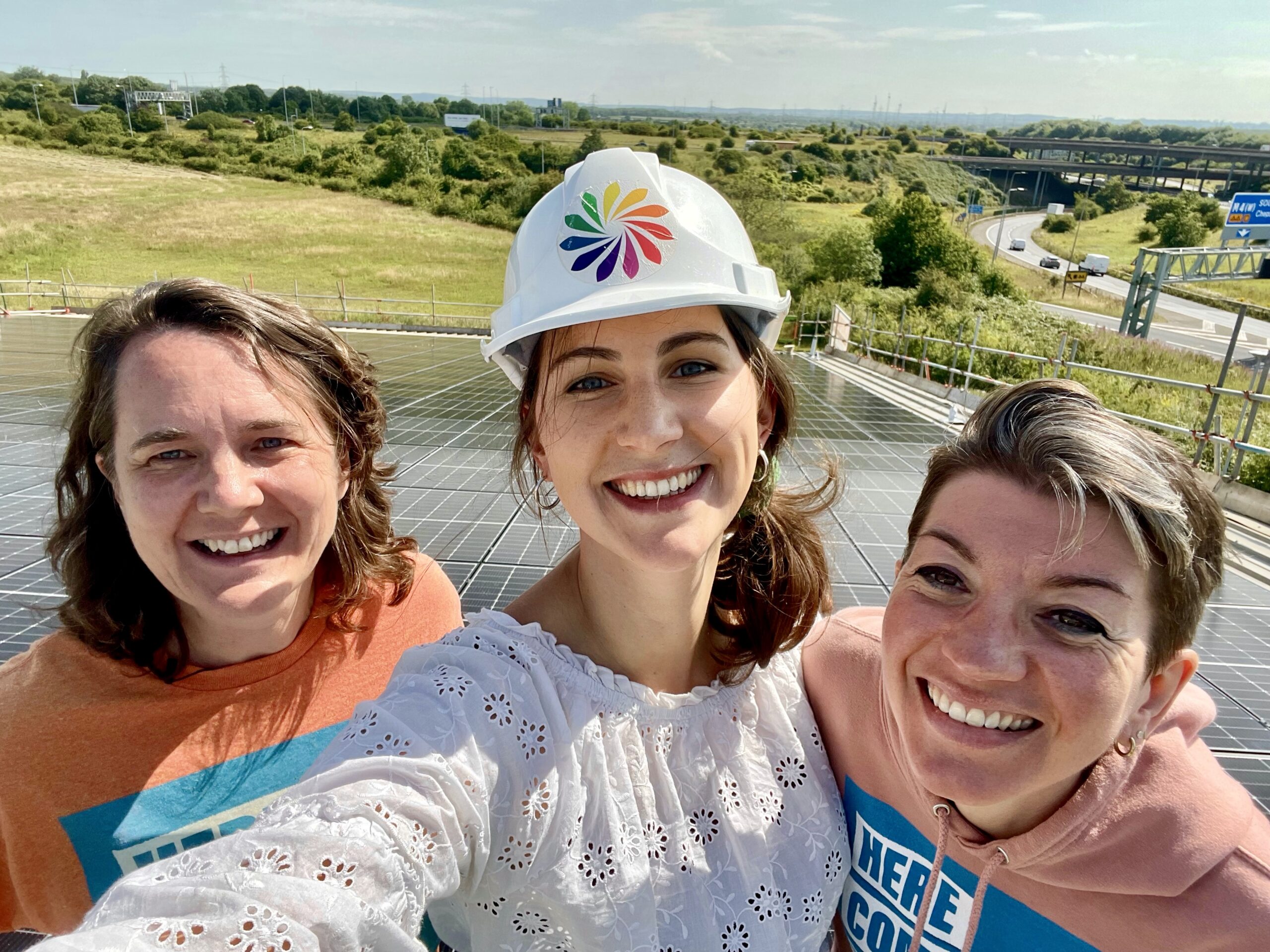Three people, one wearing a hard hat, smile into the camera in front of a solar panels and a motorway