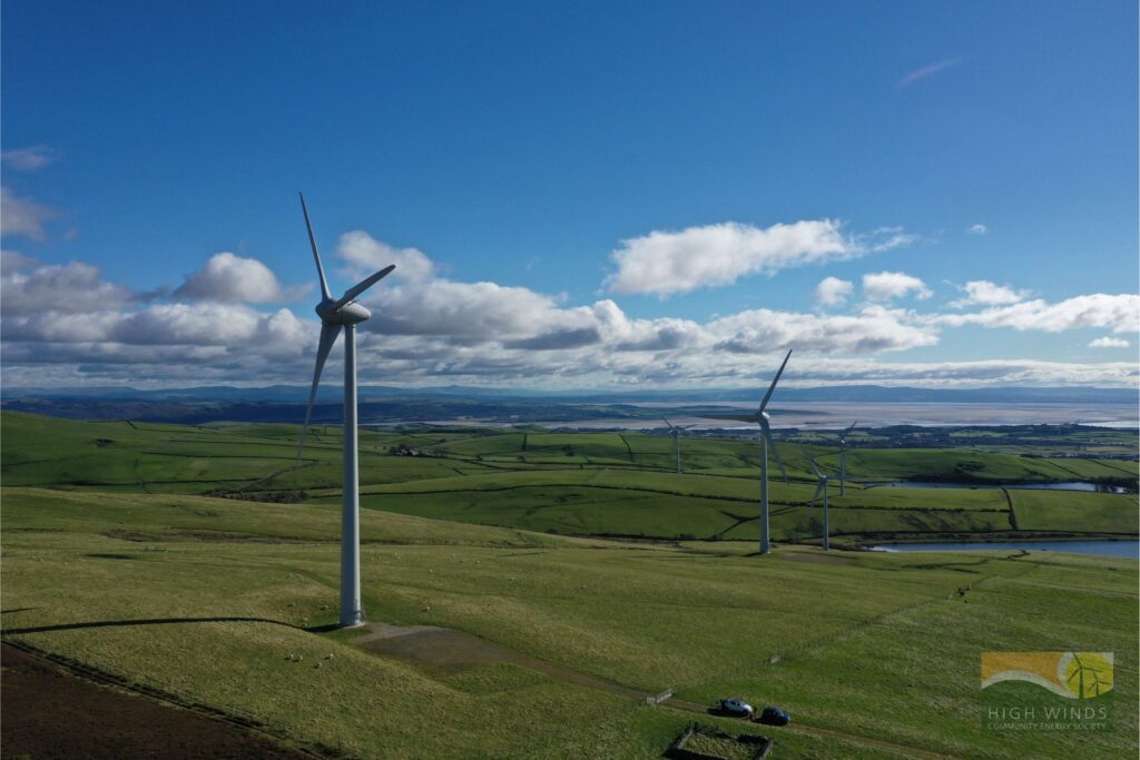 Image taken from the air of a number of wind turbines in green fields against blue skies with clouds