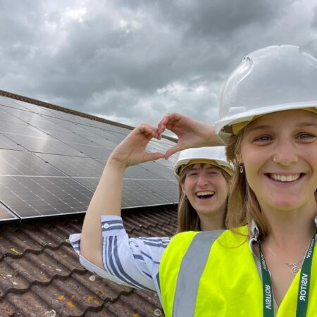two smiling people in hard hats stand on a roof next to solar panels, making a heart shape with their hands