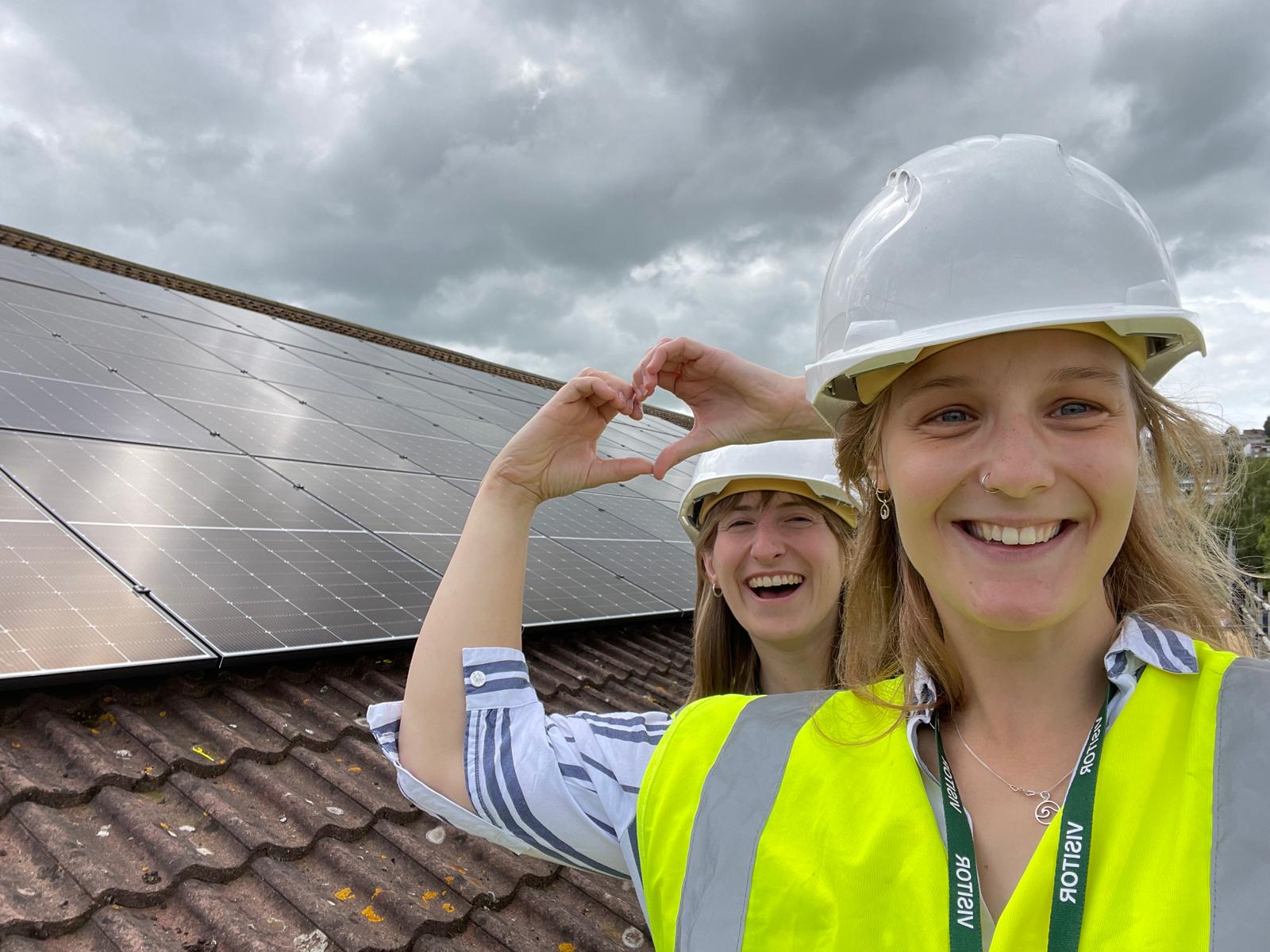 two smiling people in hard hats stand on a roof next to solar panels, making a heart shape with their hands