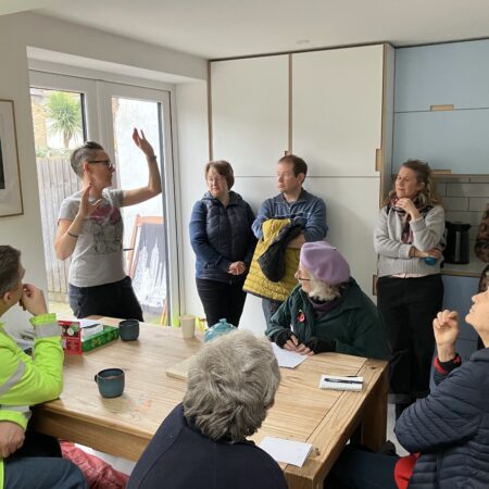 people stand and sit in a kitchen, listening to another person who is speaking and gesticulating