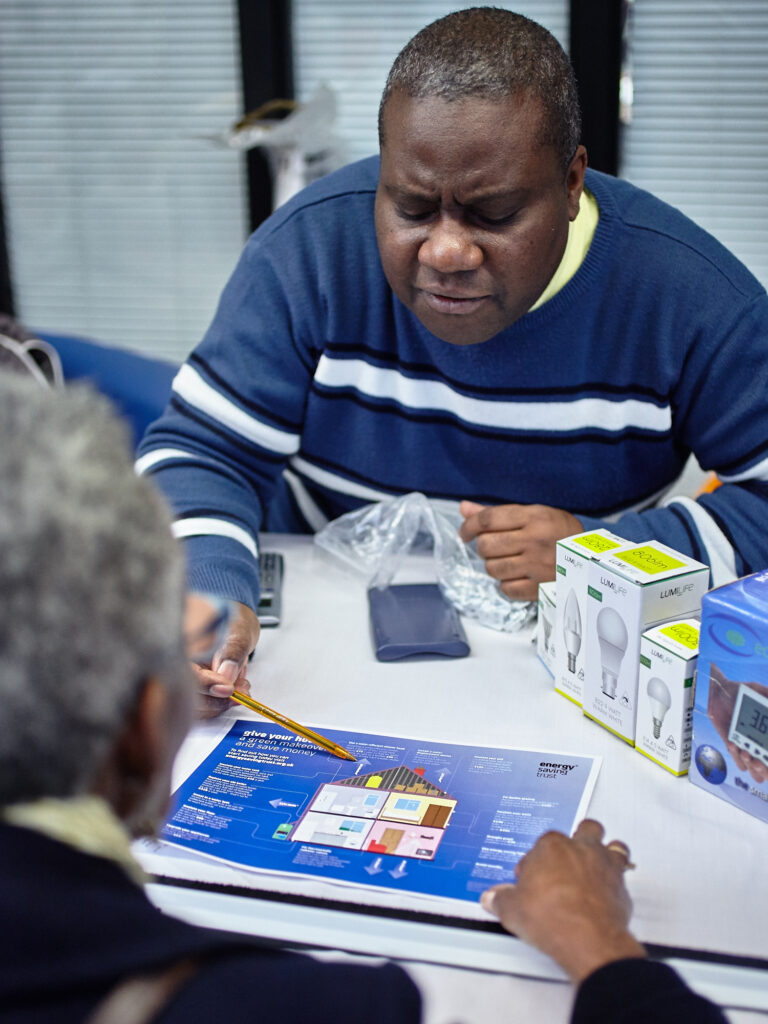 a person uses a pencil to point to a part of an illustration of a house, speaking with a person in the foreground whose back is to the camera