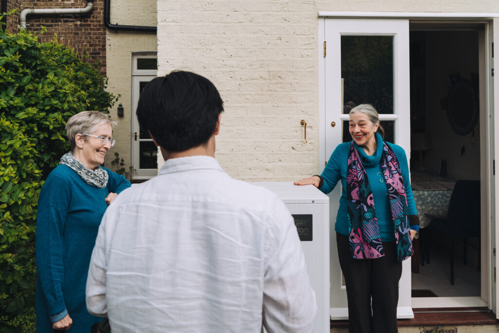 two people stand next to a heat pump, smiling at a person in the foreground whose back is to the camera