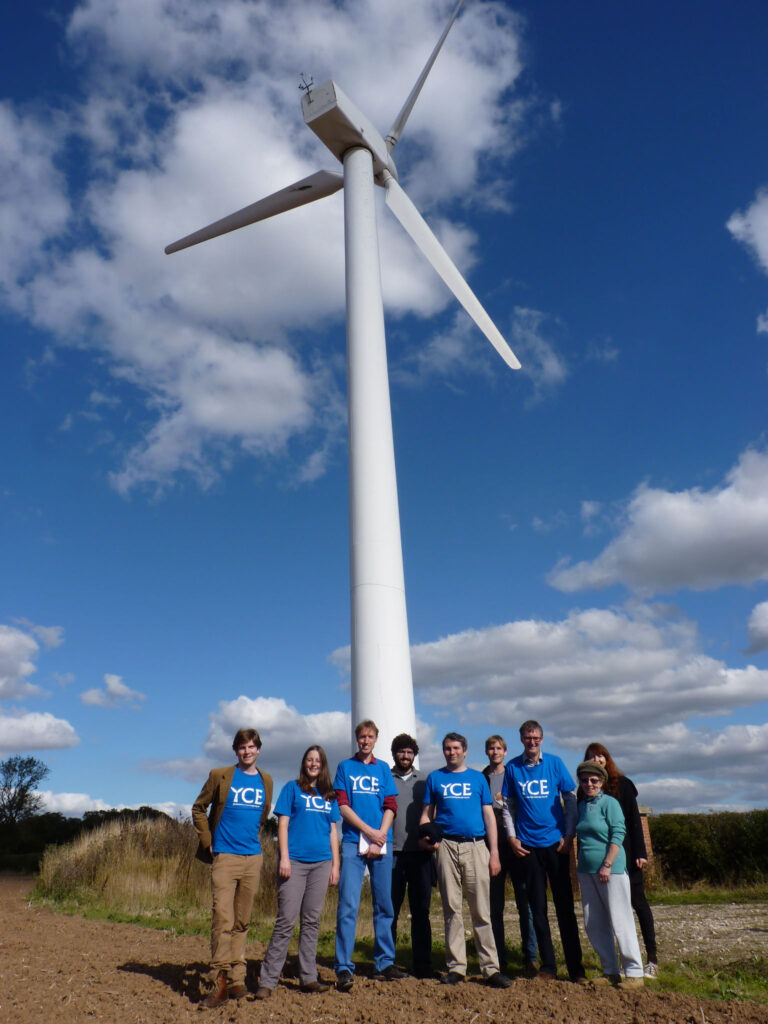 Nine people stand at the foot of a wind turbine