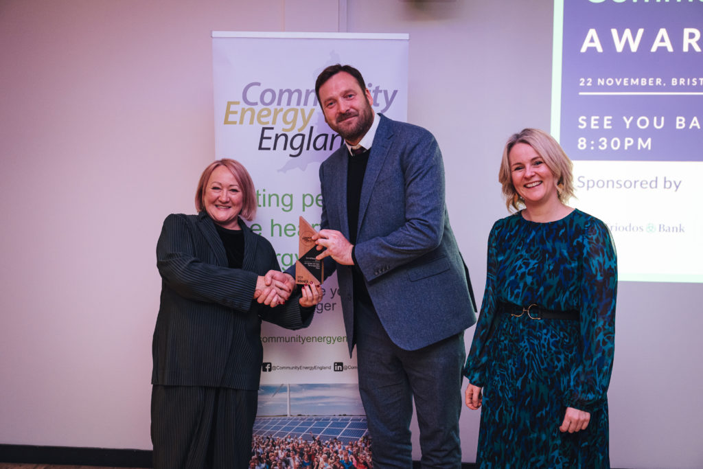 one person hands over a trophy to another while shaking their hand and a third person, standing next to the third person, while they all smile at the camera