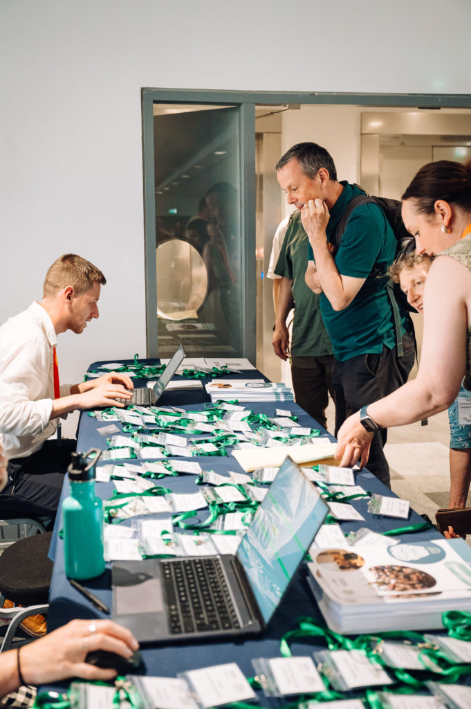 a registration desk for a conference with name badges laid out and people collecting them from a person with a laptop sitting at the table