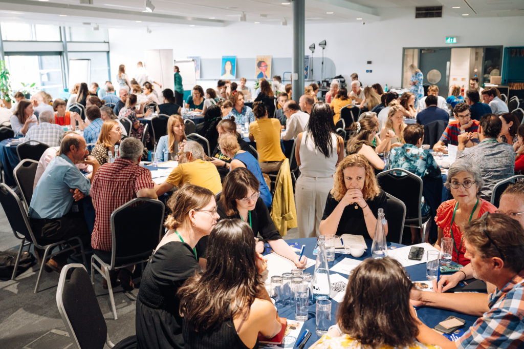a busy conference with people sat around multiple round tables, having conversations