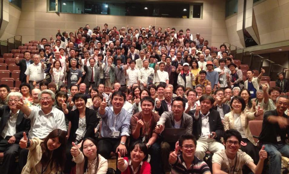 a large group of people smile at the camera, many of whom are holding thumbs up