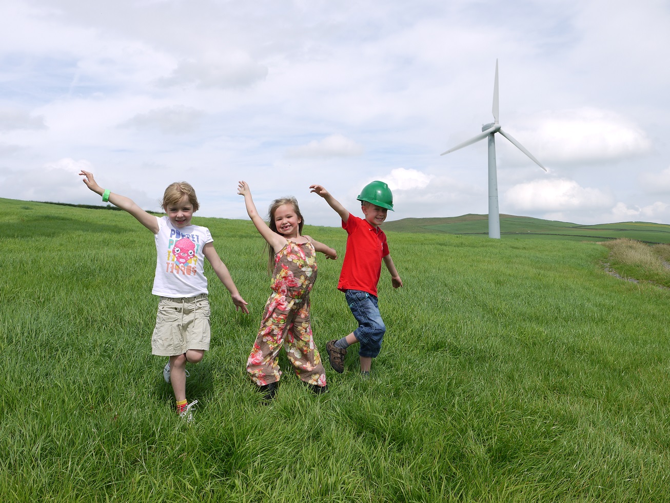 Three kids play in a grassy field with a wind turbine in the background