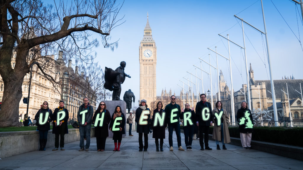 a group of people wearing lit up letters spelling out 'Up the Energy' stand in front of Big Ben and whitehall