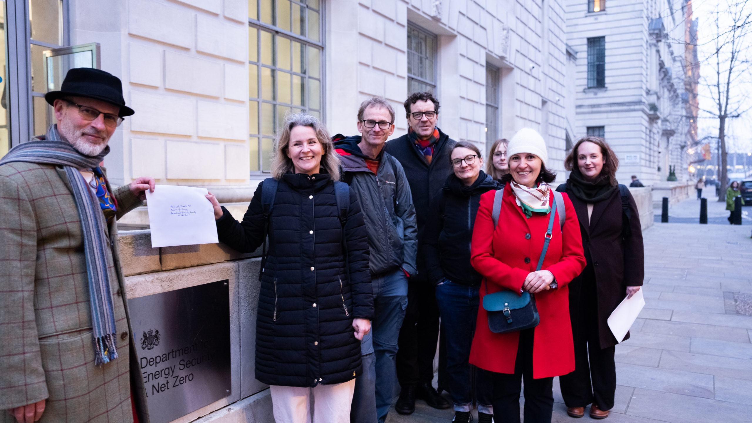 A group of eight community energy advocates standing outside the Department for Energy Security & Net Zero in London on 4 March 2026, delivering the "Up the Energy" open letter. On the far left, Duncan Law (Head of Policy & Advocacy at Community Energy England) and Emma Bridge (CEO of Community Energy England) hold the letter beside the department's sign. The remaining group members, from left to right, are: Andy O'Brien (Co-founder, Bristol Energy Cooperative), Matt Vickers (Emma's successor as CEO), Jessica Vallentine (COO of Community Energy England), Katherine Linsley (Community Energy London), Monika Paplaczyk (Thrive Renewables), and Kim Woodcock (Communications & Events Officer at Community Energy England). The group is smiling, dressed in winter coats, with the stone-fronted government building behind them.