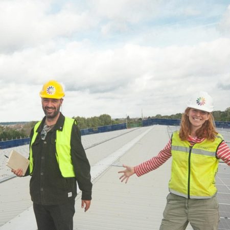 Two people in hard hats and high-visibility vests smiling on a large rooftop solar installation, with rows of solar panels stretching into the distance.