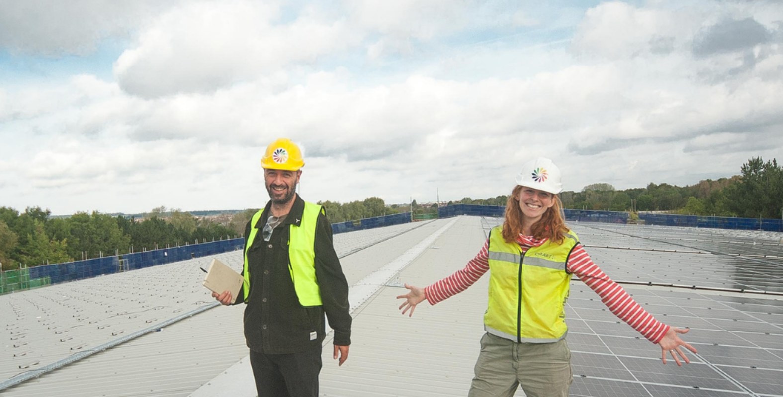 Two people in hard hats and high-visibility vests smiling on a large rooftop solar installation, with rows of solar panels stretching into the distance.